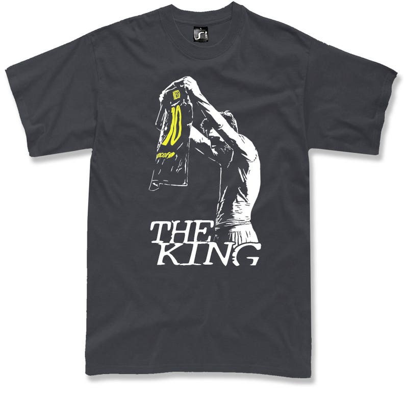 May include: Dark gray t-shirt featuring a white graphic of a person holding up a jersey with the number 10 in yellow. The text "THE KING" is printed in white below the graphic. The shirt is a crew neck.