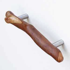 May include: A wooden cabinet pull with a natural brown finish and a unique, curved shape. The pull is attached to two silver metal brackets.