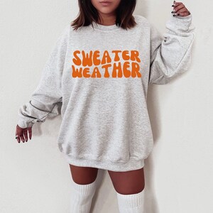 Puede incluir: Una sudadera gris con el texto "SWEATER WEATHER" en letras naranjas de estilo retro.