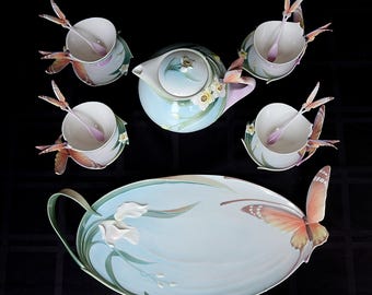 Pode incluir: Um conjunto de chá em porcelana composto por um bule, quatro chávenas com pires, quatro colheres e um tabuleiro oval. Cada peça é decorada com tons de azul pastel, detalhes florais brancos e motivos de borboletas tridimensionais.