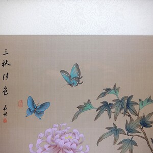 Puede incluir: Pintura de estilo asi&aacute;tico con dos mariposas azules, un crisantemo rosa y hojas verdes. La obra incluye caracteres chinos y un sello rojo sobre un fondo beige.