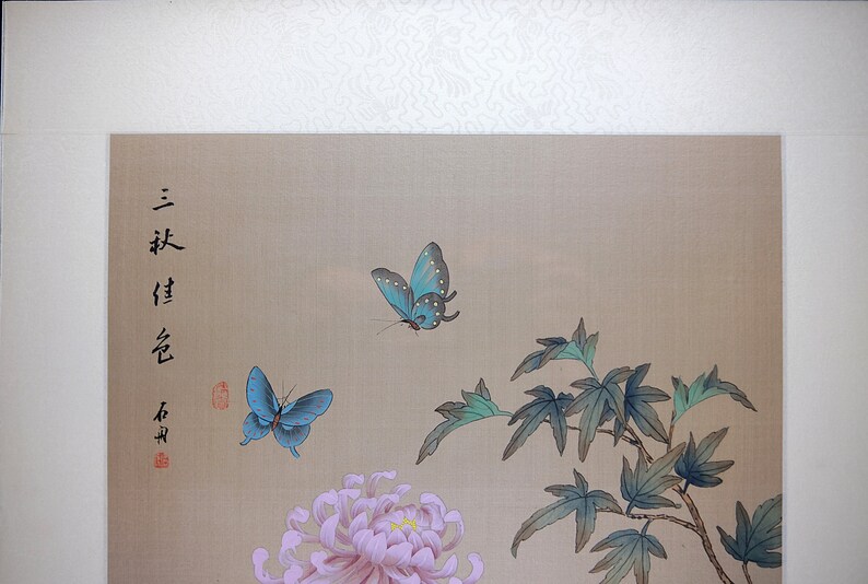 Puede incluir: Pintura de estilo asi&aacute;tico con dos mariposas azules, un crisantemo rosa y hojas verdes. La obra incluye caracteres chinos y un sello rojo sobre un fondo beige.