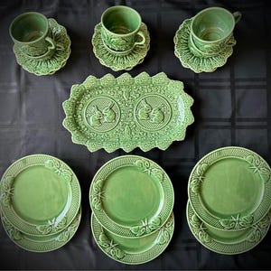 May include: A sage green ceramic dinnerware set featuring three cups with matching saucers, three plates, and one rectangular serving tray. Each piece displays embossed rabbit and floral motifs with scalloped edges and a glossy finish.