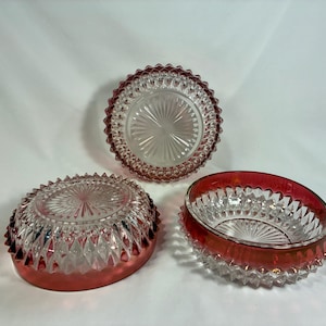 May include: Three vintage glass bowls with a ruby red trim. The bowls have a clear, textured design with a starburst pattern in the center. The bowls are in different shapes and sizes, perfect for serving or display.