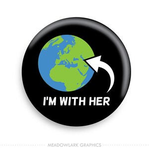 May include: A black button with a glossy finish. The button features a green and blue graphic of the Earth with a white arrow pointing towards it. The text "I'M WITH HER" is displayed in white.