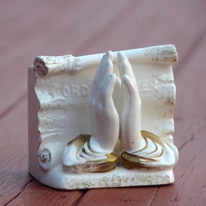 May include: White ceramic figurine of two hands clasped in prayer, with a scroll behind them that reads "The Lord's Prayer".
