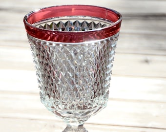 Puede incluir: Una copa de vidrio transparente con un patrón de diamante y un borde rojo. La copa está sobre una superficie de madera.