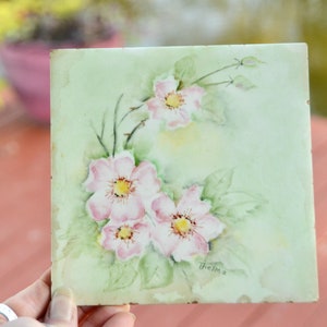 Könnte beinhalten: Quadratische, handbemalte Fliese mit rosa Blüten mit gelben Zentren und grünen Blättern auf hellgrünem Hintergrund. Die Fliese ist mit "Thelma" signiert. Die Fliese wird von einer Hand gehalten.