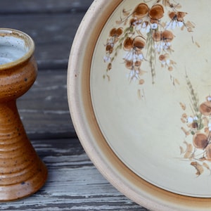 May include: A handmade ceramic goblet and a large serving plate. The goblet is brown with a speckled texture and a flared base. The plate has a cream-colored interior with a floral design in brown and white, and a brown rim.