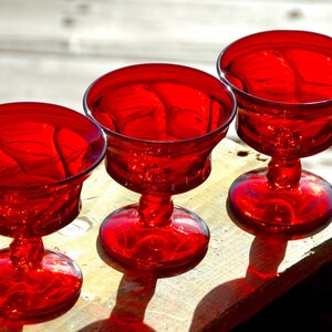 May include: Three red glass dessert cups with a swirl design. The cups are on a wooden surface.