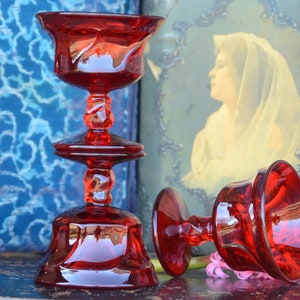 May include: Two red glass candle holders with a twisted stem and a wide base. The candle holders are on a dark wooden surface.