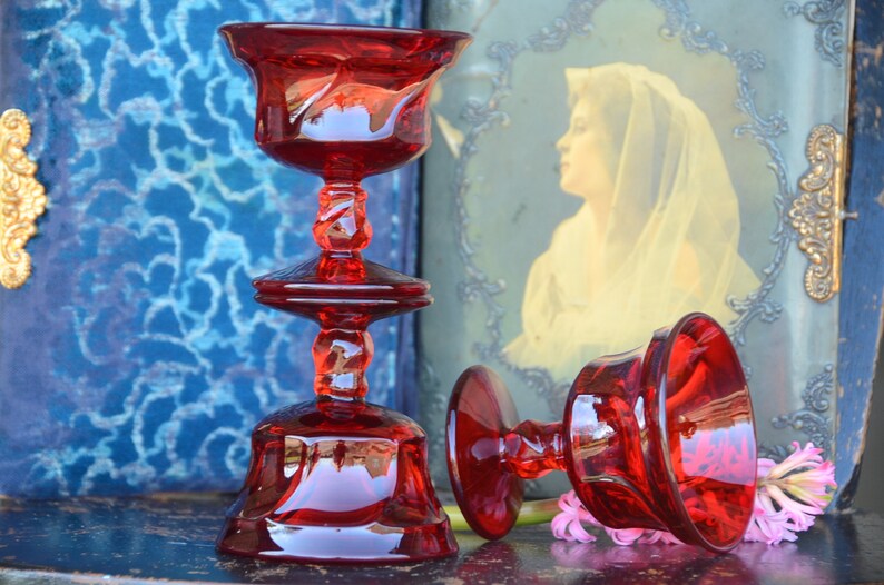 May include: Two red glass candle holders with a twisted stem and a wide base. The candle holders are on a dark wooden surface.