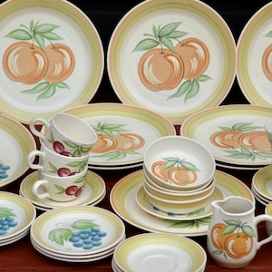 May include: A collection of ceramic dinnerware featuring fruit designs. The set includes plates, bowls, cups, saucers, and a creamer. The plates and bowls have a yellow border and are decorated with peaches, grapes, and cherries.