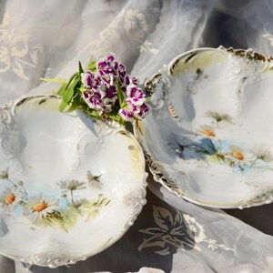May include: Two white porcelain bowls with gold trim and floral designs. The bowls have a scalloped edge and are decorated with delicate flowers and leaves. The bowls are sitting on a white fabric background.