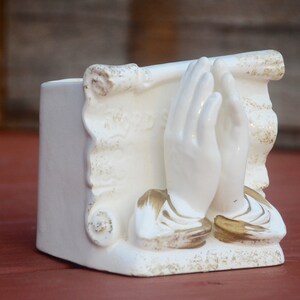 May include: White ceramic planter shaped like a pair of hands clasped in prayer. The hands are decorated with gold accents and the planter has a scroll design.