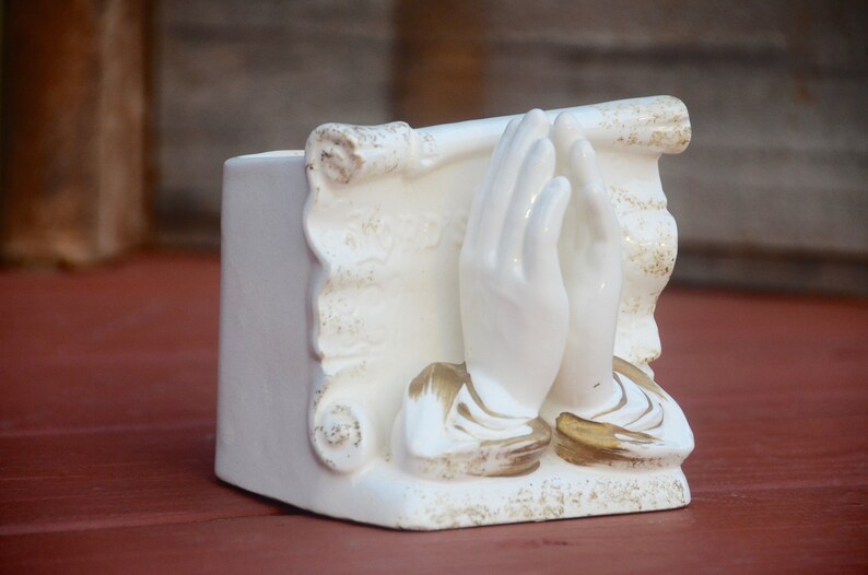 May include: White ceramic planter shaped like a pair of hands clasped in prayer. The hands are decorated with gold accents and the planter has a scroll design.