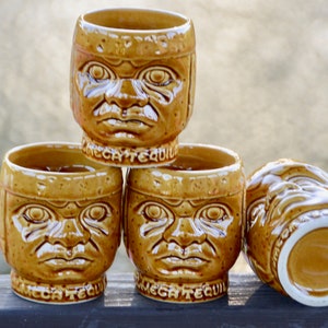 Op de afbeelding: Set van vier bruine keramische shotglazen in de vorm van gezichten met de woorden "Mega Tequila" op de bodem.