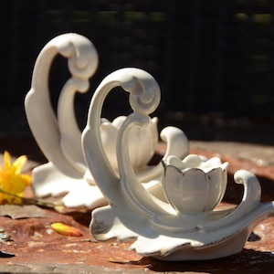 May include: Two white ceramic candle holders with a floral design. The holders are shaped like a stylised flower with a curved stem.