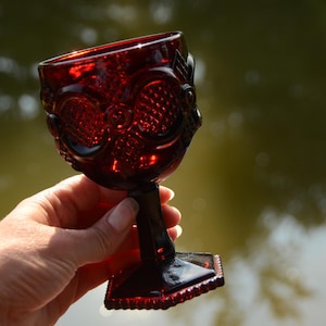 Op de afbeelding: Een diep rode glazen kelk met een decoratief patroon. De kelk heeft een ronde kom en een steel met een vierkante voet.