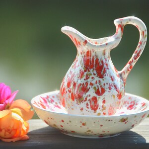 May include: A white ceramic pitcher and matching saucer with a red and gold splatter pattern. The pitcher has a handle and a rounded body. The saucer is round and has a slightly raised edge.