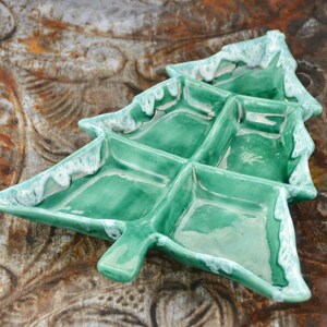 Pode incluir: Uma travessa verde de cerâmica em forma de árvore de Natal com seis compartimentos. As bordas são adornadas com uma cobertura branca texturizada. A travessa está sobre uma superfície castanha texturizada.