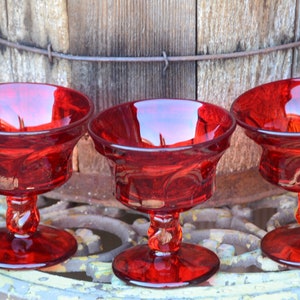 May include: Three red glass dessert dishes with a swirl design on the stem. The dishes are sitting on a metal surface.