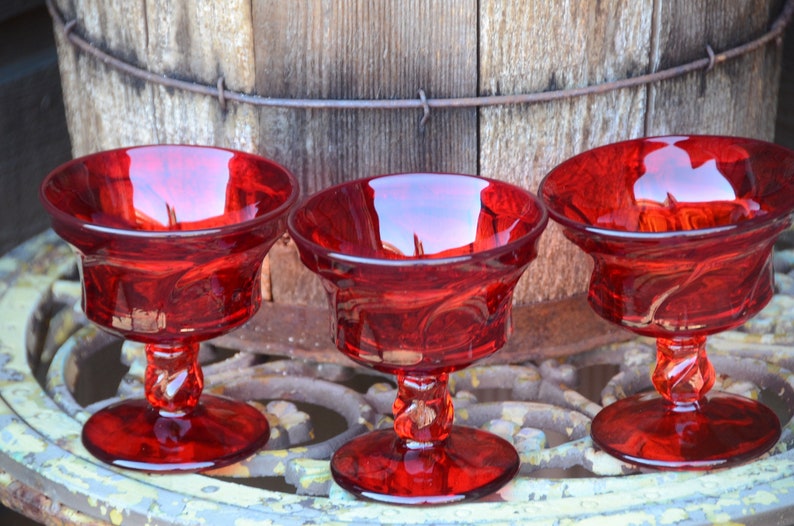 May include: Three red glass dessert dishes with a swirl design on the stem. The dishes are sitting on a metal surface.