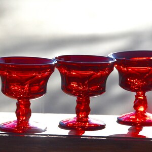 May include: Three red glass stemmed dessert cups or sherbet glasses. The glasses have a fluted design and are made of ruby red glass.