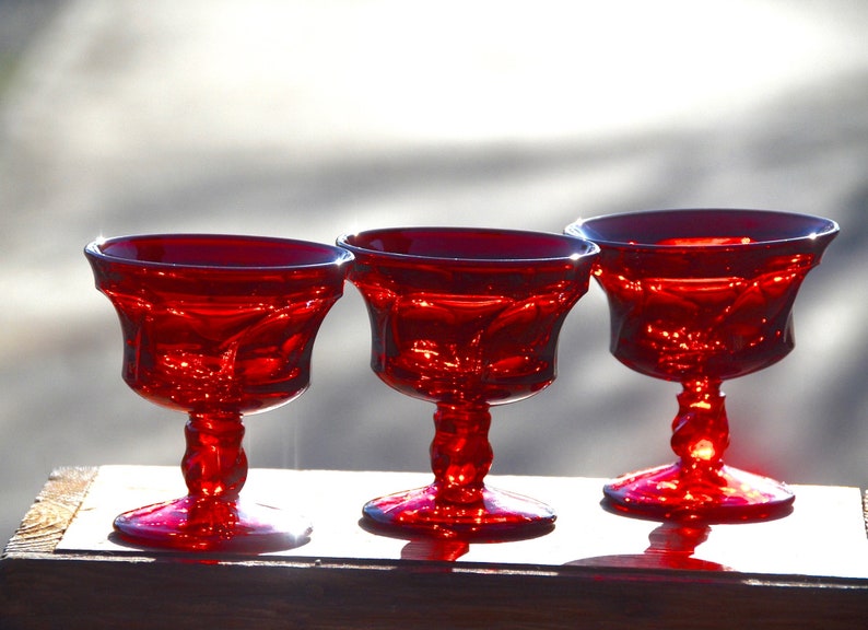 May include: Three red glass stemmed dessert cups or sherbet glasses. The glasses have a fluted design and are made of ruby red glass.