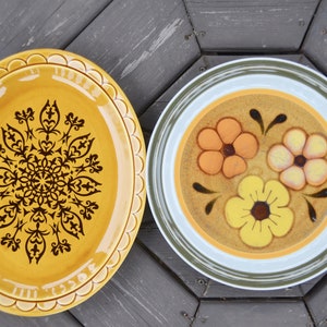 Puede incluir: Dos platos de cerámica con diferentes diseños florales. El primer plato es amarillo con un patrón geométrico marrón. El segundo plato es marrón con un borde blanco y tres flores amarillas, naranjas y rosas.
