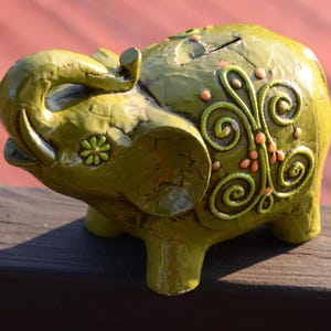 May include: A green ceramic elephant piggy bank with a floral design. The elephant has a curled trunk and a small flower on its side.