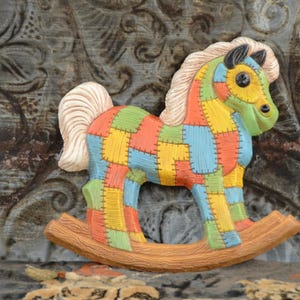 Könnte beinhalten: Ein buntes Patchwork-Schaukelpferd aus Holz, das in Blautönen, Grün, Gelb und Orange bemalt ist. Das Pferd hat eine weiße Mähne und einen weißen Schweif.