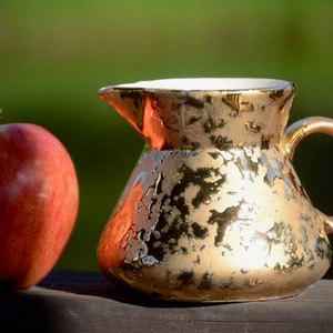 Peut inclure: Une crémier en céramique de couleur dorée avec une surface texturée et une poignée. Une pomme rouge se trouve à côté de la crémier sur une surface en bois.