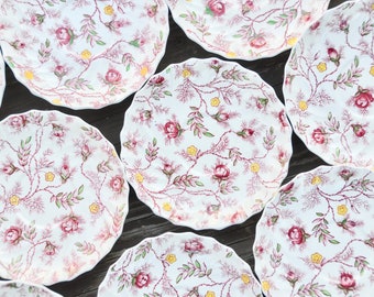 Pode incluir: Um conjunto de oito pratos de cerâmica branca com um padrão floral que apresenta rosas cor-de-rosa, flores amarelas e folhas verdes sobre um fundo branco.