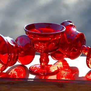 May include: Four red glass cups with a stemmed base. The cups are arranged on a wooden surface. The cups are clear and have a textured surface.