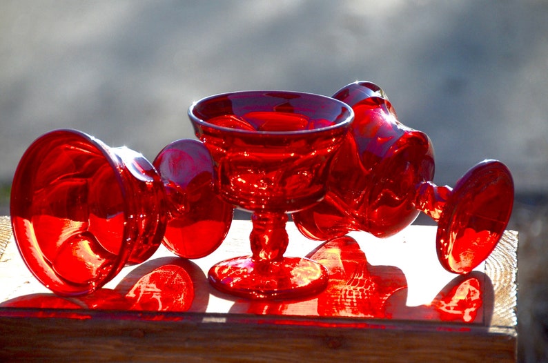 May include: Four red glass cups with a stemmed base. The cups are arranged on a wooden surface. The cups are clear and have a textured surface.