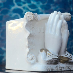 May include: White ceramic figurine of two hands clasped in prayer, with gold accents. The hands are holding a scroll with the word "Jesus" written on it.