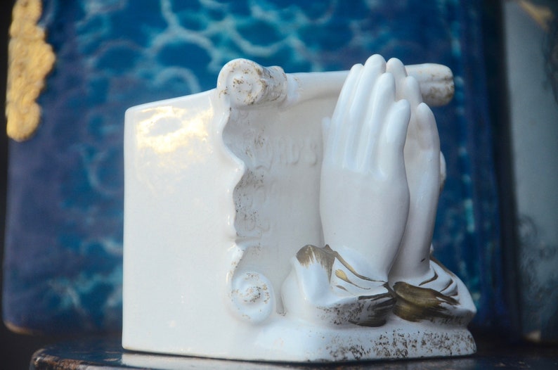 May include: White ceramic figurine of two hands clasped in prayer, with gold accents. The hands are holding a scroll with the word "Jesus" written on it.