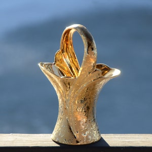 May include: A gold-colored ceramic vase with a decorative handle in the shape of a basket. The vase has a textured surface and a unique, flowing design.
