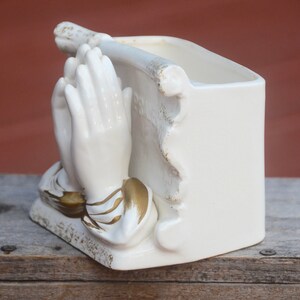 May include: White ceramic planter shaped like a pair of hands clasped in prayer. The hands are detailed with gold accents. The planter has a rectangular opening for planting.