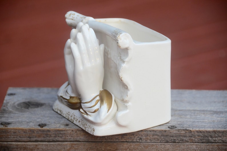 May include: White ceramic planter shaped like a pair of hands clasped in prayer. The hands are detailed with gold accents. The planter has a rectangular opening for planting.