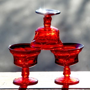 May include: Three red glass pedestal dishes, one stacked on top of the other. The top dish is a small, round bowl with a wide, flat base. The two bottom dishes are larger and have a more traditional goblet shape.
