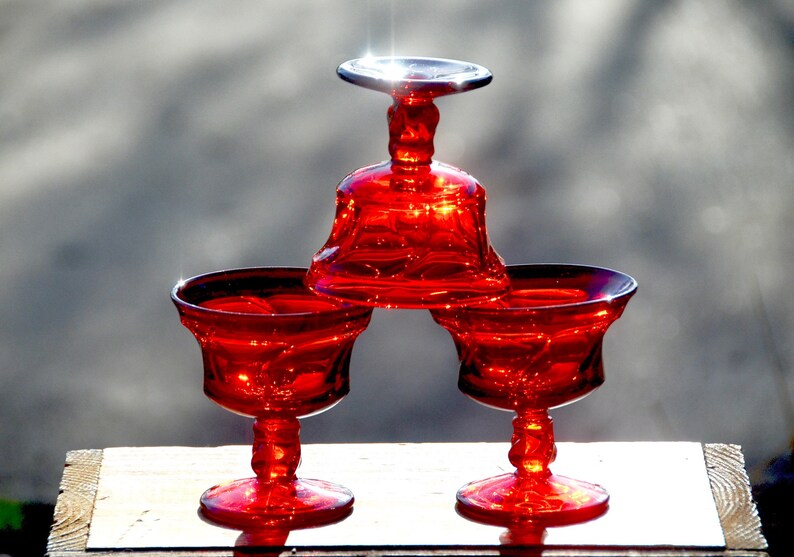 May include: Three red glass pedestal dishes, one stacked on top of the other. The top dish is a small, round bowl with a wide, flat base. The two bottom dishes are larger and have a more traditional goblet shape.