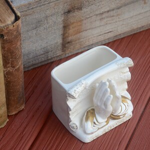 May include: White ceramic planter shaped like two hands clasped in prayer. The planter has gold accents and is sitting on a red wooden surface.