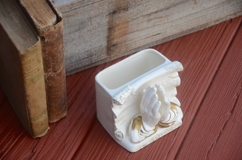 May include: White ceramic planter shaped like two hands clasped in prayer. The planter has gold accents and is sitting on a red wooden surface.