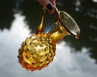 May include: A amber glass pitcher with a textured, bubbled surface and a wide, flared spout. The pitcher has a handle and is partially filled with liquid.