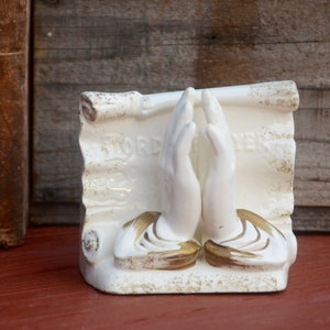 May include: White ceramic bookends featuring two hands clasped in prayer with gold accents. The hands are positioned on top of a scroll with the word "LORD" and "PRAYER" written on it.