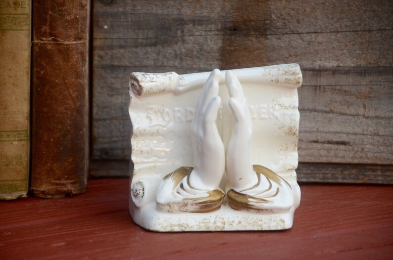 May include: White ceramic bookends featuring two hands clasped in prayer with gold accents. The hands are positioned on top of a scroll with the word "LORD" and "PRAYER" written on it.