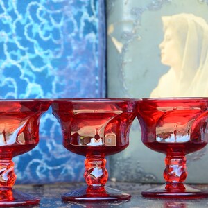 May include: Three red glass stemmed cups with a swirl design. The cups are arranged in a row on a dark surface.