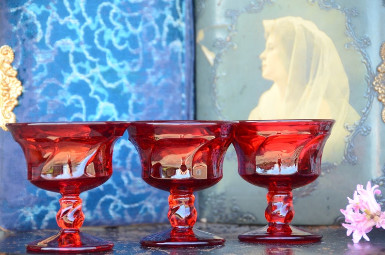 May include: Three red glass stemmed cups with a swirl design. The cups are arranged in a row on a dark surface.